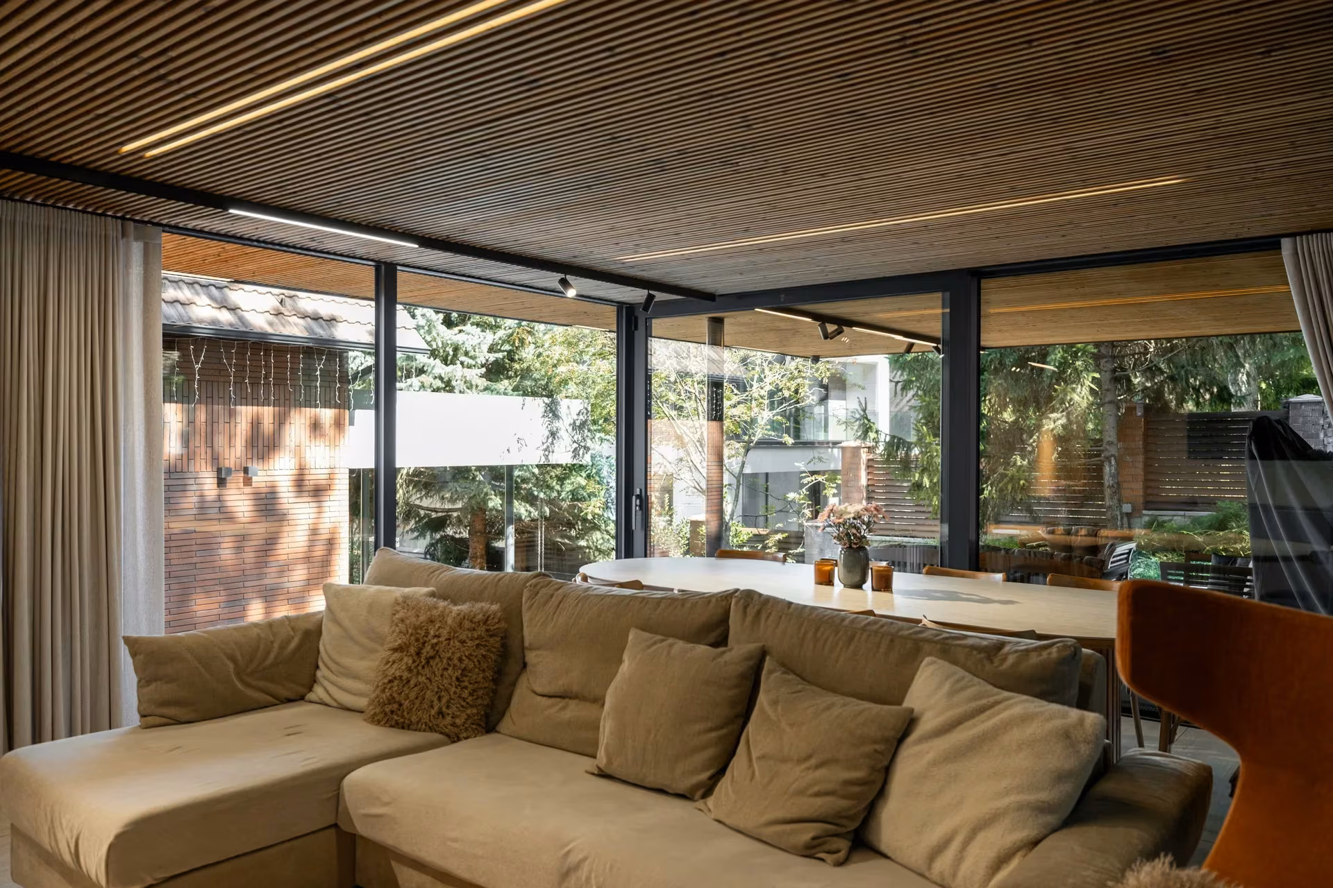 Living room with a light-coloured sofa, timber ceiling and panoramic glazing overlooking the garden.