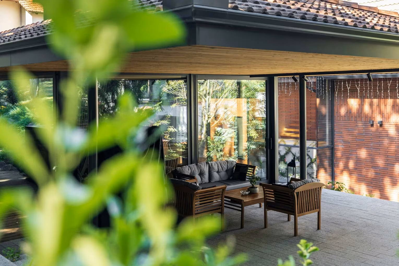 Covered terrace with timber furniture and expansive glazing, photographed through garden foliage.