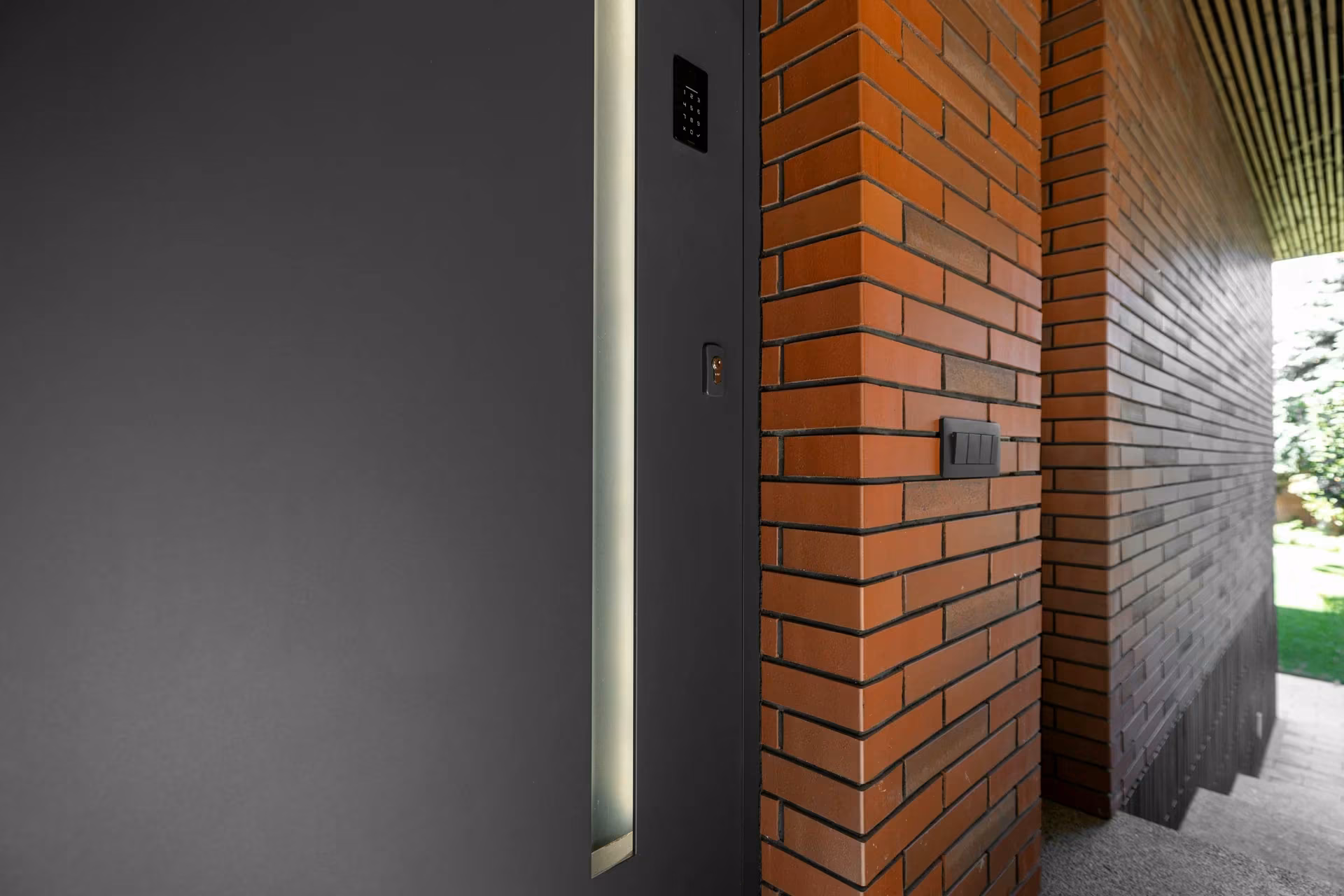 Detail of a dark entrance door beside brickwork and a wall-mounted access element.