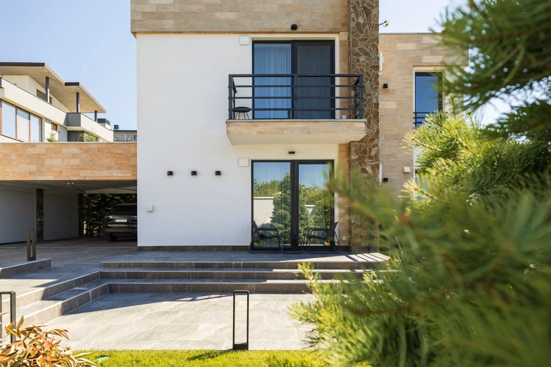 Side facade of the house with panoramic windows, stone-textured cladding and nearby greenery.